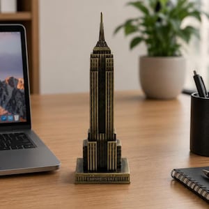 May include: A bronze-coloured metal model of the Empire State Building featuring a stepped architectural design and a pointed spire. The base of the structure is rectangular and clearly displays the embossed text EMPIRE STATE.