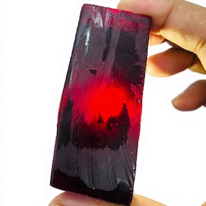 Pode incluir: Uma gema de rubi bruta, retangular, com um tom vermelho profundo. A pedra tem uma superfície áspera e texturizada com áreas de coloração escura, quase preta. O rubi é segurado contra um fundo branco, destacando sua cor vibrante e forma natural.