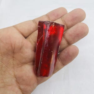 May include: A translucent, deep red gemstone with a rough, uneven surface. The rectangular shape of the stone is visible, with light passing through the material. The gemstone is held in a hand against a white background.