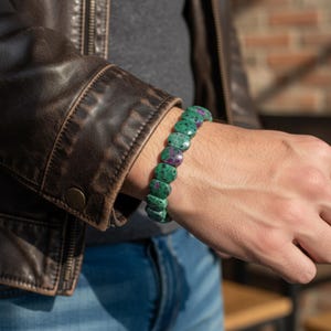 Puede incluir: Un brazalete de piedras preciosas verdes y rojas. El brazalete está hecho de piedras ovaladas en tonos verdes y rojos. El brazalete se lleva en la muñeca, con una chaqueta de cuero marrón en el fondo.