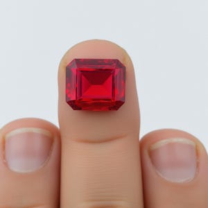 May include: A faceted, rectangular-cut, red gemstone. The gem is held between two fingers against a white background. The gem has a square shape with beveled edges and a brilliant, reflective surface.