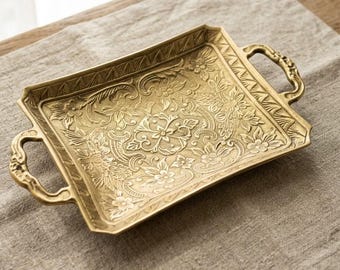Puede incluir: Una bandeja rectangular de latón con un intrincado patrón floral en relieve en toda su superficie. La bandeja incluye dos asas curvas y ornamentadas en los lados opuestos y un borde decorativo con un diseño geométrico repetido.