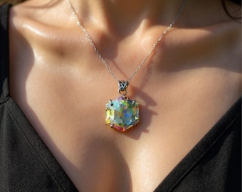 May include: A silver necklace with a large, iridescent, hexagonal pendant. The pendant reflects multiple colors, including yellow, green, and pink. The necklace is worn against a dark background.