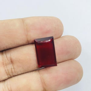 Op de afbeelding: Een rechthoekige, dieprode edelsteen met een gefacetteerde bovenkant. De edelsteen wordt in een hand gehouden tegen een witte achtergrond. De kleur en slijpvorm van de steen suggereren dat het een robijn of granaat zou kunnen zijn.