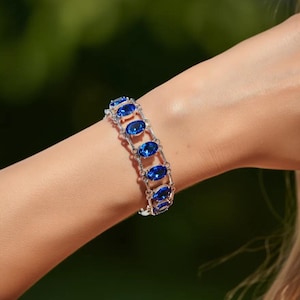 Puede incluir: Un brazalete plateado con piedras preciosas azules ovaladas. El brazalete está compuesto por una serie de eslabones unidos, cada uno con una piedra preciosa. Las piedras preciosas están engastadas en metal plateado con un diseño delicado y ornamentado. El brazalete se lleva en la muñeca.