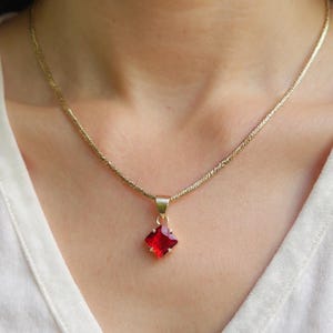 May include: A gold-coloured necklace featuring a square, red gemstone pendant. The pendant is a vibrant red and set in gold-coloured metal. The necklace chain has a textured design.