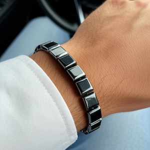 Puede incluir: Una pulsera gris oscuro con eslabones cuadrados se lleva en la muñeca. La pulsera está hecha de un material metálico brillante. La persona lleva una camisa blanca y vaqueros azules.