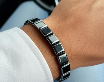May include: A dark gray bracelet with square links is worn on a wrist. The bracelet is made of a shiny, metallic material. The person is wearing a white shirt and blue jeans.