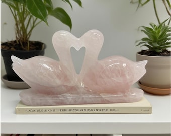 May include: A pink rose quartz sculpture of two swans with their necks forming a heart shape. The sculpture sits on a book and is displayed on a white shelf. The swans are a light pink colour.