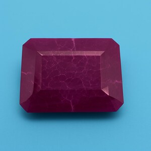 May include: A rectangular, faceted gemstone in a deep magenta colour. The gem features a network of fine, light-coloured veins across its surface. The gemstone is set against a solid, light blue background.