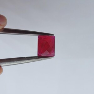 以下が含まれることがあります： ピンセットで挟まれた、長方形のファセットルビーの宝石。宝石は鮮やかで深い赤色で、研磨された表面を持っています。石のカットは複数の内部反射を生み出し、その輝きを増しています。