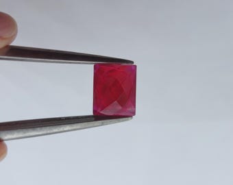 May include: A faceted, rectangular ruby gemstone held by tweezers. The gem is a vibrant, deep red color with a polished surface. The cut of the stone creates multiple internal reflections, enhancing its brilliance. The background is a plain, light gray.