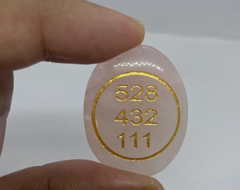 Puede incluir: Una piedra lisa de cuarzo rosa de forma ovalada con un diseño circular dorado. El diseño presenta los números 528, 432 y 111 en color dorado. La piedra está sujeta en una mano.