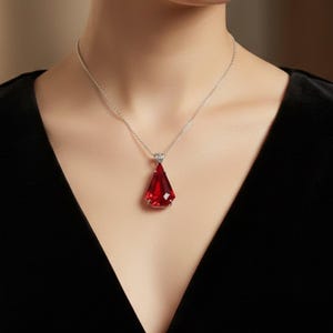 May include: A silver necklace featuring a faceted, teardrop-shaped, red pendant. The pendant hangs from a delicate chain. The necklace is set against a black background. A piece of jewellery.