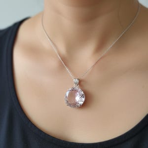 May include: A silver necklace featuring a large, round, pale pink gemstone pendant. The pendant is suspended from a delicate silver chain. The necklace is worn against a dark background.