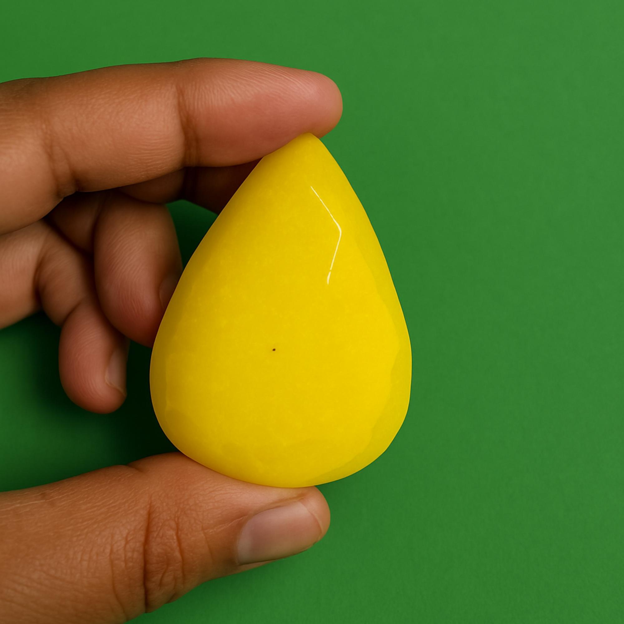 May include: A bright yellow, teardrop-shaped gemstone. The polished stone is held against a green background. The gem has a smooth, glossy surface and a faceted design. The gemstone is a vibrant, solid color.