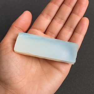 May include: A rectangular, translucent stone with a pale blue and white gradient. The stone is held in a hand, showcasing its smooth surface and natural, raw edges. The stone's color transitions from white to a soft blue.