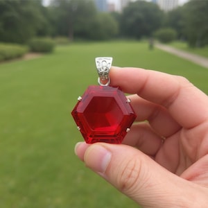 May include: A hexagonal, faceted red pendant with a silver-coloured bail. The pendant is held against a blurred green background, suggesting an outdoor setting. The gemstone-like pendant is a striking piece of jewellery.