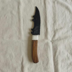 May include: A handcrafted knife featuring a dark, curved blade and a wooden handle. The blade has a distinctive, notched pattern. The handle is wrapped with white material near the blade. The knife is presented on a white cloth.