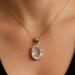 Op de afbeelding: Een zilveren ketting met een heldere, ovale hanger. De hanger is gezet in een zilveren bezel en hangt aan een fijne zilveren ketting. De ketting wordt getoond tegen een donkere achtergrond.