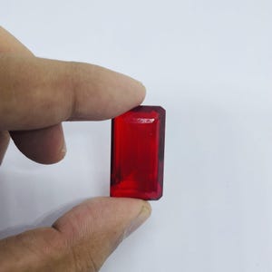 Peut inclure: Une pierre précieuse rouge foncé, rectangulaire et à facettes, est tenue entre deux doigts sur un fond blanc. La gemme a une surface polie et un aspect clair et transparent. La gemme est une pierre semblable à un rubis.