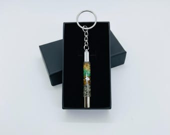 May include: A silver keychain with a cylindrical pendant filled with colorful stones and a silver cap. The keychain is presented in a black gift box against a white background. The pendant contains green, white, and brown stones.
