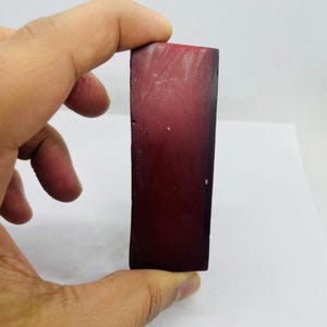 Puede incluir: Una piedra rectangular, de color rojo oscuro, sostenida entre los dedos. La piedra tiene una superficie lisa y pulida y un color profundo y rico. La forma es un bloque simple y alargado, con una textura ligeramente desigual. La piedra mide unos 7,6 cm de largo.