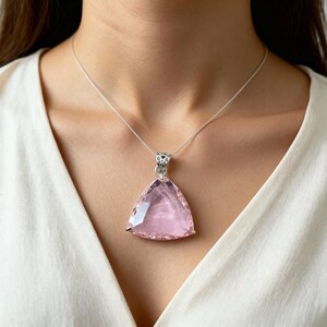 May include: A silver necklace featuring a large, triangular, pink gemstone pendant. The pendant is a soft pink color and has a faceted surface. The necklace is worn against a white top, creating a contrast.