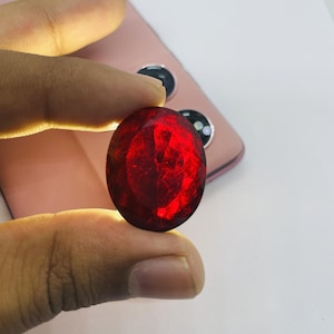 May include: A large, oval-shaped, deep red gemstone with multiple facets. The gem is held between fingers, with a pink smartphone in the background. The stone's facets reflect light, creating a brilliant display of colour.
