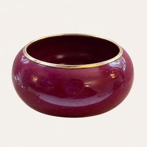 May include: A deep magenta bangle bracelet with a smooth, glossy finish and a thin, metallic gold-toned rim along the top edge. The circular accessory features a rounded, bulbous profile and a reflective surface that catches light across its curve.