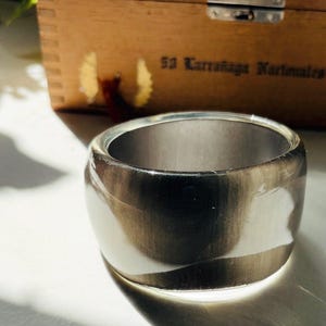 May include: A wide, circular bangle bracelet featuring a polished finish with a swirling pattern of translucent white and dark charcoal gray tones. The bracelet has a smooth, rounded exterior surface and a plain, metallic-colored interior band.