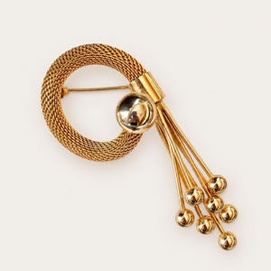 May include: A gold-tone brooch featuring a circular mesh ring attached to a smooth, polished gold-tone sphere. Five thin, dangling gold-tone chains extend from the sphere, each ending in a small, polished gold-tone bead.
