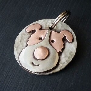 May include: A silver and copper dog tag with a cartoon dog face. The dog has a copper nose and ears, and a silver body. The tag has a silver ring for attaching to a collar.