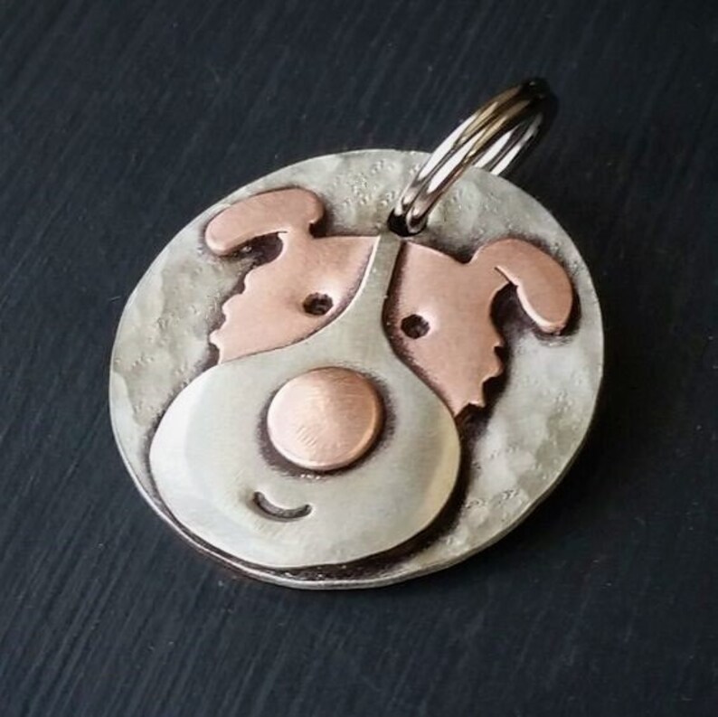 May include: A silver and copper dog tag with a cartoon dog face. The dog has a copper nose and ears, and a silver body. The tag has a silver ring for attaching to a collar.