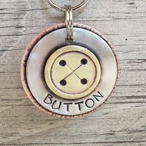 May include: A silver and copper dog tag with a brass button design. The tag has the word "BUTTON" engraved on it.