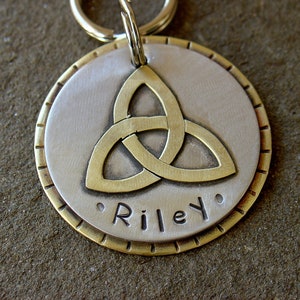 May include: A silver and brass dog tag featuring a Celtic knot design with the name "Riley" engraved on it.