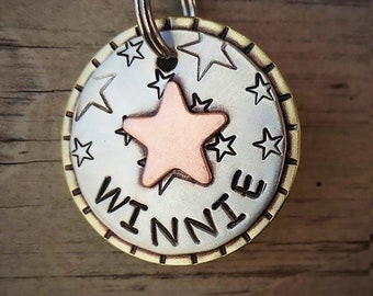 May include: A round, silver-colored pet tag with a bronze star in the center and smaller star cutouts around it. The name "WINNIE" is stamped in black letters. The tag is attached to a metal ring.