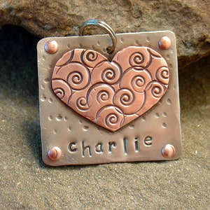 May include: A silver metal pet tag with a copper heart-shaped design featuring a swirl pattern. The tag is engraved with the name "charlie".