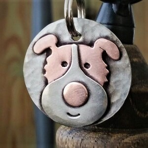 May include: A silver and copper dog-shaped tag with a smiling face. The tag has a ring for attaching to a collar or keychain.