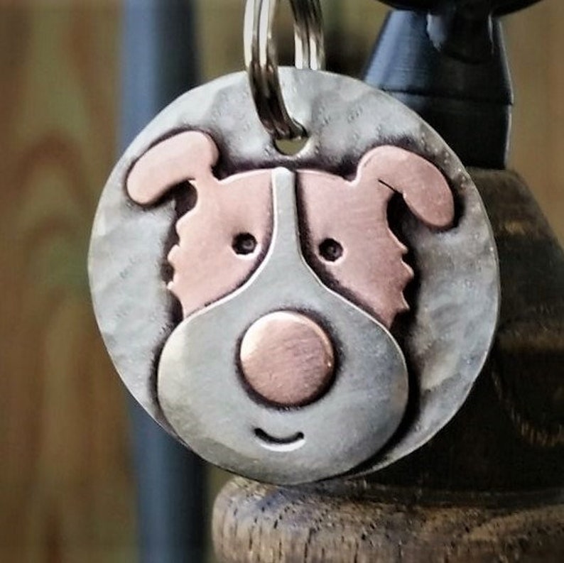 May include: A silver and copper dog-shaped tag with a smiling face. The tag has a ring for attaching to a collar or keychain.