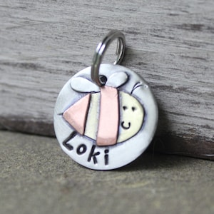 May include: A silver dog tag featuring a bee design. The bee is crafted from copper, yellow, and pink metal. The tag is engraved with the name "Loki".