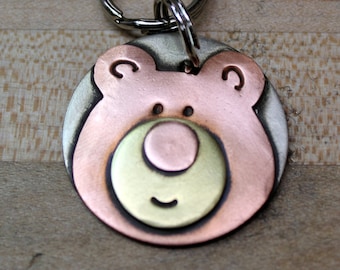 May include: A round metal keychain with a bear face design. The bear is pink with a gold nose and mouth. The keychain is attached to a silver ring.