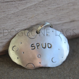 May include: Silver dog tag with a textured surface and a rounded shape. The tag has the name "Spud" engraved on it.