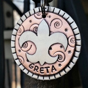 May include: Round metal pet tag with a fleur-de-lis design. The tag features a silver fleur-de-lis on a copper-colored background with swirls and star accents. The name "GRETA" is engraved at the bottom, and the tag has a silver border.