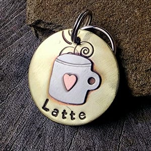 Può includere: Un medaglietta rotonda per animali domestici, color ottone, con un disegno di tazza da caffè argentata e un cuore rosa. La parola "Latte" è scritta in nero sotto la tazza. Un anello argentato è attaccato in alto.