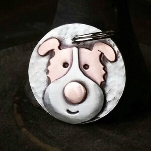 May include: A silver dog tag with a copper-colored dog face. The dog has a large, round nose and a happy expression.