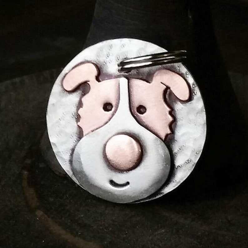 May include: A silver dog tag with a copper-colored dog face. The dog has a large, round nose and a happy expression.