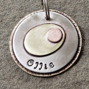 May include: A layered metal pendant with a silver, brass, and copper design. The pendant has an oval shape in the center and the name "Ollie" is engraved at the bottom. A silver keyring is attached at the top.