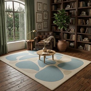 May include: A living room with a patterned rug. The rug has a light beige base with large, irregular shapes in light blue and beige. The room also features a leather armchair, a round coffee table, and a wooden bookcase filled with books.