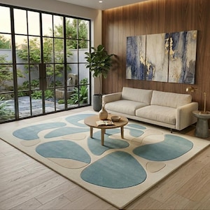 May include: A contemporary living room with a large rug featuring abstract blue and off-white shapes on a light beige background. A light-coloured sofa, round wooden coffee table, and artwork are present. A large window offers a view of a garden.
